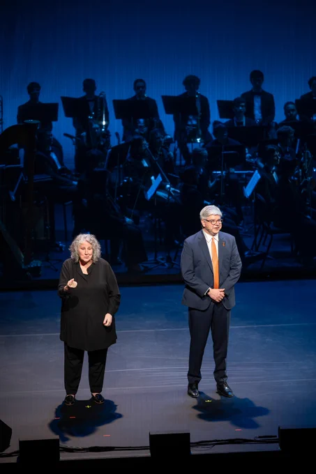 An American Sign Language interpreter stands on stage next to the R I T president Bill Sanders. An orchestra is behind them 