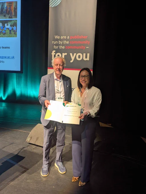 Two individuals standing on a stage holding an open certificate or award, with a backdrop displaying the text 'We are a publisher run by the community for the community for you' in a professional event setting.