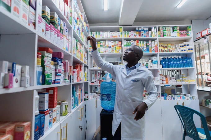 Man in lab coat in pharmacy aisle reaching for a medicine