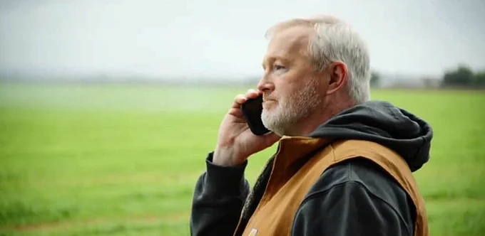 A farmer standing in a green field, talking on a cellphone, appearing thoughtful or concerned.