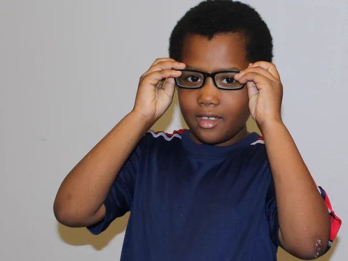 A person wearing a navy blue sports jersey adjusts glasses while looking at the camera. The background is a plain light-colored wall.