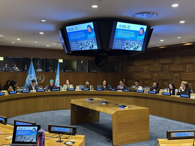 Delegates seated around a large circular conference table in a formal meeting room with United Nations flags and screens displaying presentation slides.
