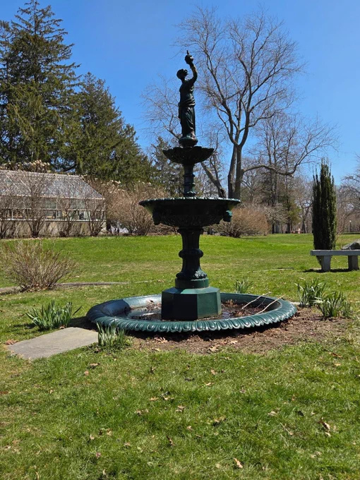 A dark green, tiered fountain topped with a small statue stands on a grassy lawn, surrounded by early spring trees under a clear blue sky at Buttonwood Park.