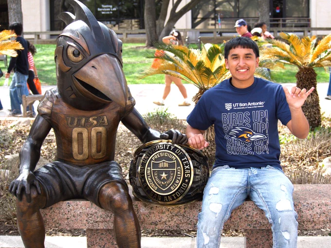 A student sits next to a bronze statue of the UT San Antonio mascot Rowdy while giving the "birds up" sign, with pinkie and thumb extended. 