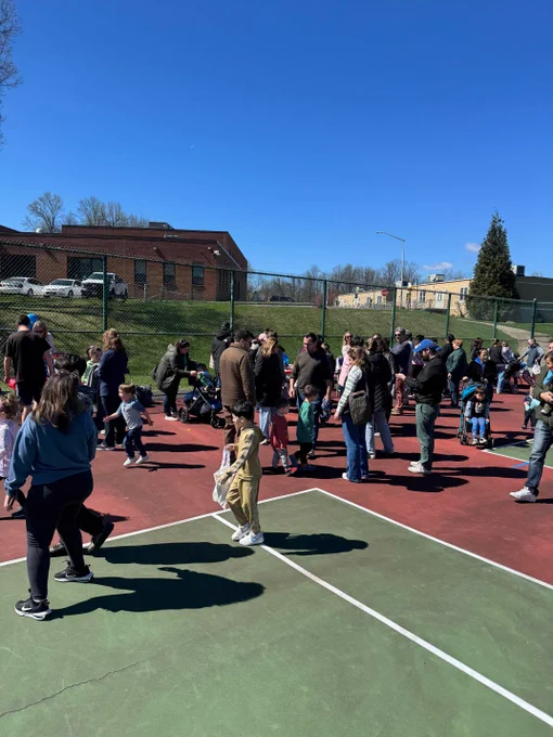 Outdoor basketball court with people playing and spectating on a sunny day.