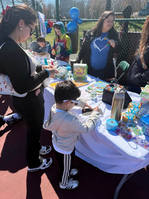 Outdoor gathering with tables set up for an event, people making crafts and decorations, colorful balloons, and a table covered with party supplies.
