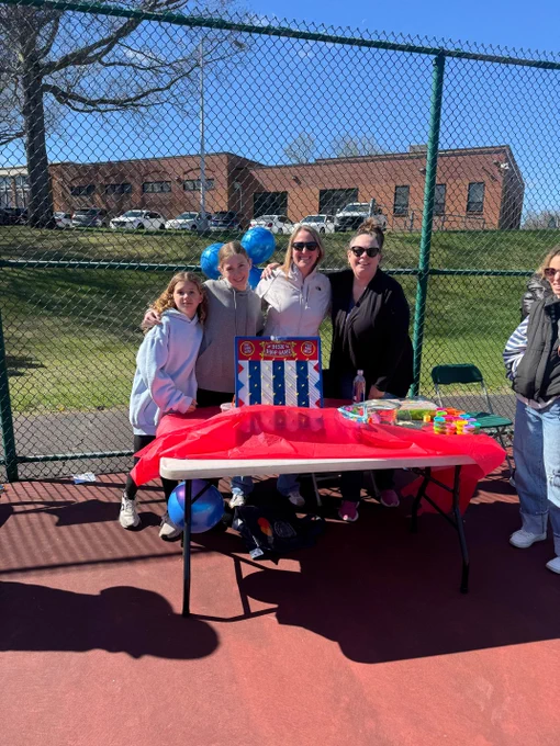 Group of people gathered around a table with red tablecloth and outdoor activities equipment on a sports field surrounded by fencing.
