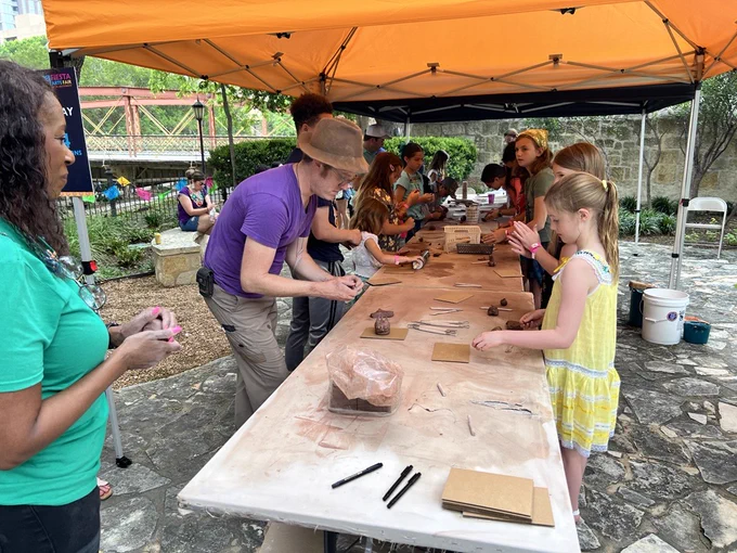 A group of people is gathered under an orange tent, engaging in a pottery activity. They are shaping clay at a long table covered with tools and materials. The setting appears to be outdoors, with trees and a stone path nearby.