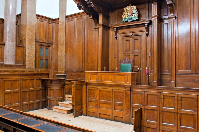 Empty courtroom with polished wooden paneling, a judge's bench, and a green leather chair beneath a royal crest.