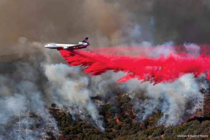 A large aircraft releases a stream of bright red fire retardant over a forested area, combating an active wildfire. Smoke rises from the burning landscape. Power lines are visible in the bottom left corner. Copyright Mark Edward Harris.