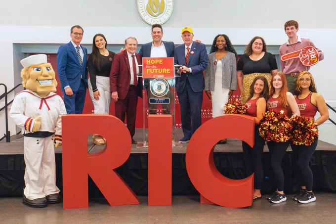 A group of people is gathered on a stage behind large red letters spelling "RIC." A mascot stands on one side, and cheerleaders with pom-poms are on the other. A "Hope for the Future" sign is displayed at the center, and there are various individuals dressed in formal and casual attire.