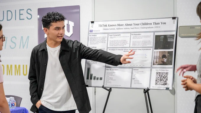 A person is standing near a presentation board titled "TikTok Knows More About Your Children Than You," gesturing while talking to others. The board includes charts, text, and a QR code. There are blurred words "STUDIES," "ISM," and "COMMUN" on a background wall.