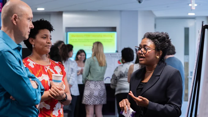 A group of people engaged in conversation at a professional event. One side displays a board with text related to mentoring programs. The setting is indoors with a focus on networking and discussion.