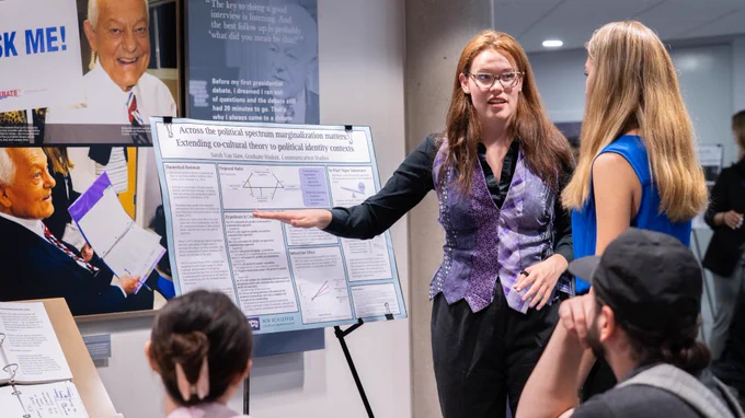 A person presents a research poster to three people in a room. The poster discusses political spectrum and marginalized status with visible charts and text. A notable photo of Bob Schieffer is visible in the background.