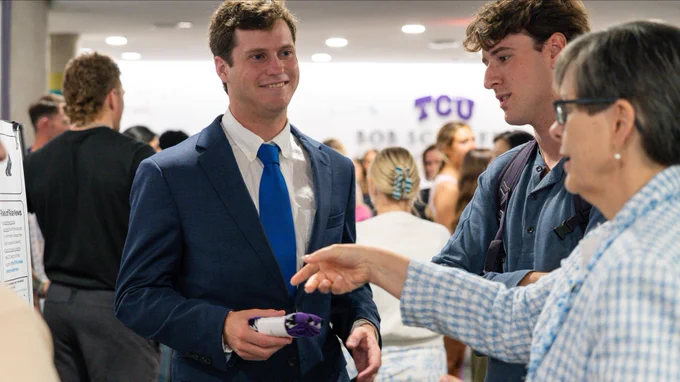 A person in a suit holding a purple item engages in conversation with others at an event. In the background, a TCU sign is visible, indicating a university setting. Attendees are mingling and exchanging information.