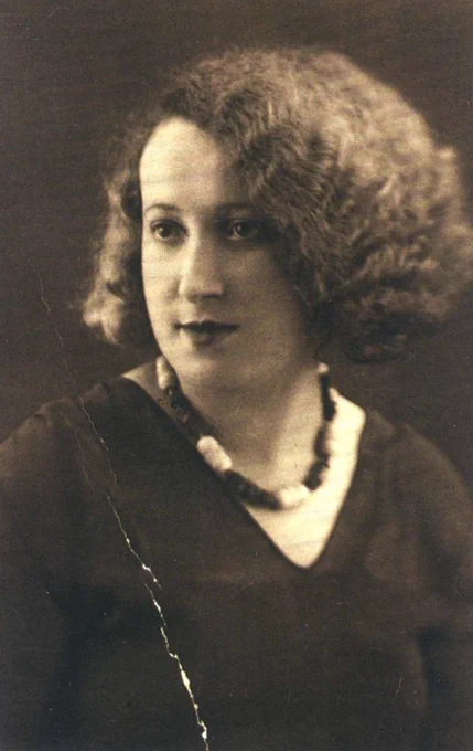 Portrait photograph of a woman. She has very wavy hair. It falls mainly on the left side of her head. She is wearing beads and a blouse with a V-cut neckline. 