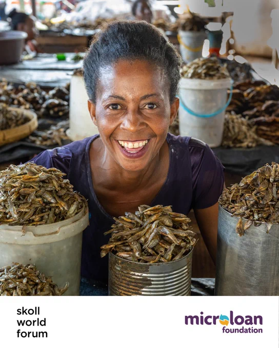 A close-up photograph of a woman smiling broadly behind a market stall. In front of her are several large tin cans and plastic buckets filled to the brim with small, dried fish. The background shows a busy, blurred market setting. The bottom of the image is white with the "Skoll World Forum" logo on the left and the "MicroLoan Foundation" logo on the right.