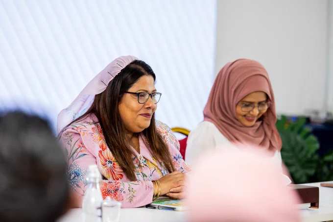 Minister Geela Ali smiling, PS is seen in the background also smiling. Both are sitting down