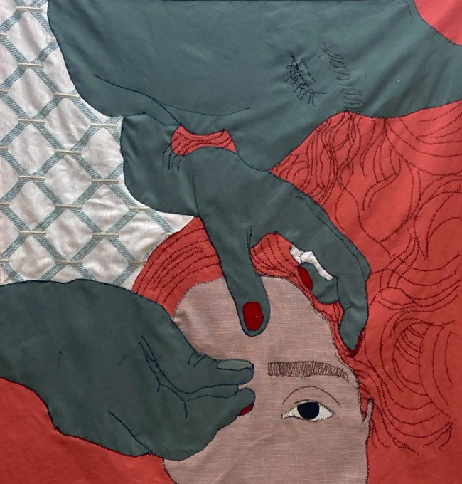 Lightly-wrinkled textile portrait: color-blocked fabrics pieced together with zigzag-stitch line work. A dark green figure with red nails carefully threads the thick brows of a redhead with voluminous hair. A quilt-like pattern fills the background.