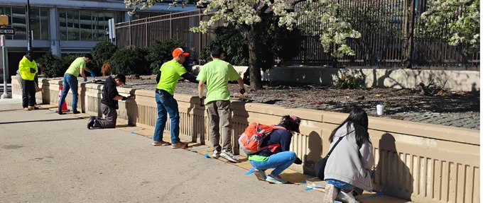 Group of people painting a concrete wall outdoors on a sunny day, wearing casual clothes and safety vests.