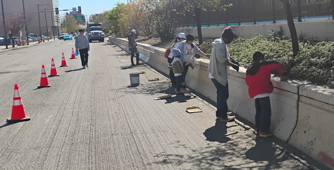 Volunteers cleaning and painting a concrete barrier along a city street lined with traffic cones on a sunny day.
