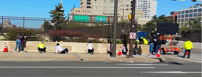 Group of people painting a concrete barrier along a city street on a sunny day.