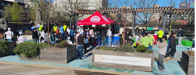 Volunteers gathered around a red tent with a logo in an outdoor park area on a sunny day.