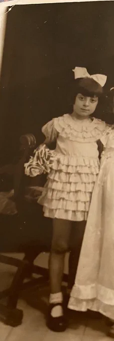 A girl standing at a table (visible part of it to the right of the frame). She is wearing a dress with elongated ruffles. A large white bow is tied on her head. 