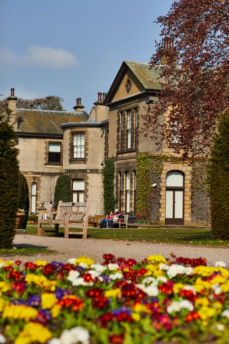 A view of Lotherton Hall from an angle, capturing flowerbeds in the foreground, bushes and trees. In the distance, visitors can be seen sitting on benches.