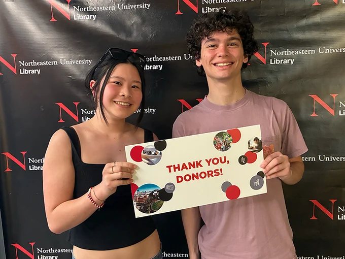 Two people smile while holding a sign that says "Thank you, donors!" They are standing in front of a black backdrop with the Northeastern University Library logo
