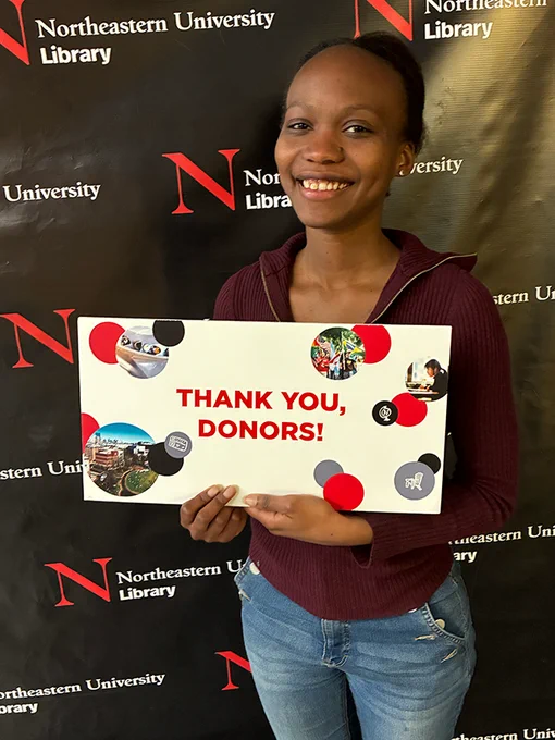 A person smiles while holding a sign that says "Thank you, donors!" They are standing in front of a black backdrop with the Northeastern University Library logo