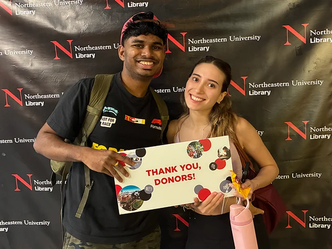 Two people smile while holding a sign that says "Thank you, donors!" They are standing in front of a black backdrop with the Northeastern University Library logo