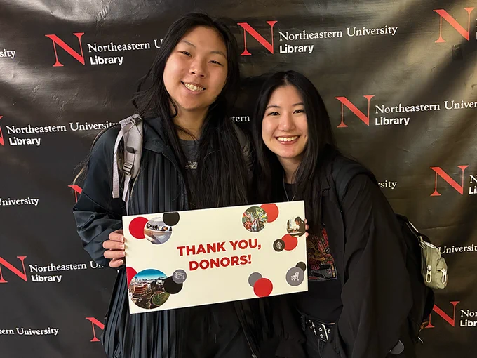 Two people smile while holding a sign that says "Thank you, donors!" They are standing in front of a black backdrop with the Northeastern University Library logo