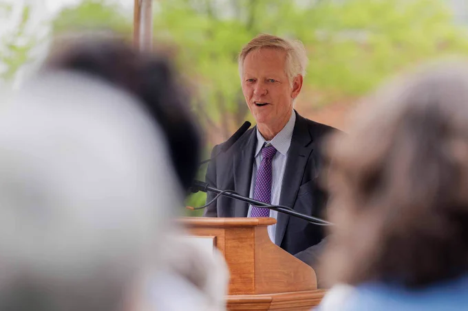 UVA President Beardsley speaking at a wooden podium outdoors, with a microphone. Greenery is visible in the background, and attendees are out of focus in the foreground.