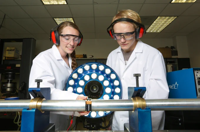 Two students wearing lab coats and safety goggles examine a device in a laboratory setting.