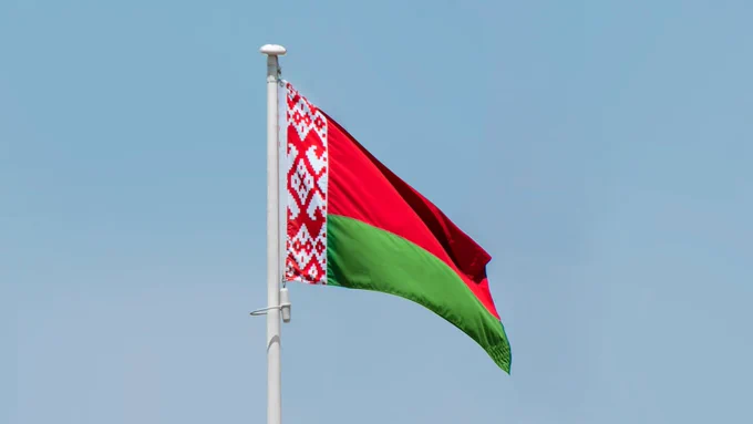 The flag of Belarus flying from a pole against a blue sky background.