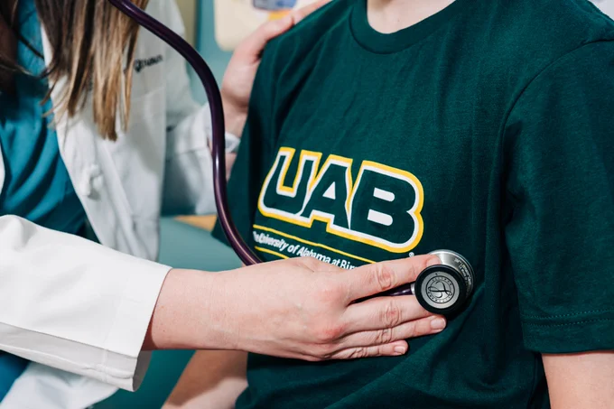 A healthcare professional uses a stethoscope to listen to the chest of a person wearing a green UAB T-shirt.