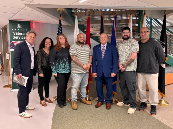 Group of seven people standing indoors near flags and a Veteran Services sign, dressed in casual and formal attire.