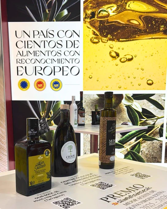 Botellas de aceite de oliva expuestas sobre mesa, con cartel sobre alimentos con reconocimiento europeo y fondo de olivas.