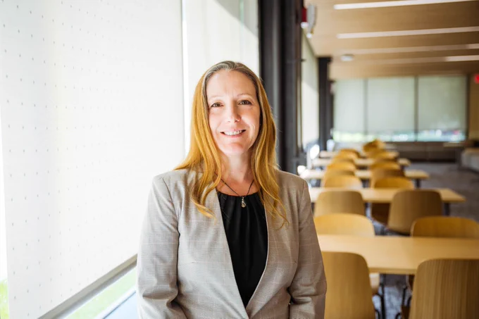 Professional woman in a gray blazer standing in a bright, modern office with wooden tables and chairs.