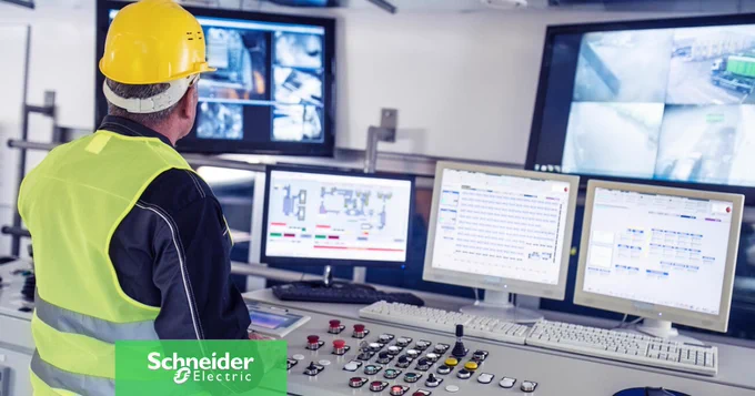 Ein Techniker mit gelbem Schutzhelm und Sicherheitsweste steht in einem Kontrollraum und \u00FCberwacht mehrere Computerbildschirme, auf denen Daten und \u00DCberwachungsaufnahmen angezeigt werden.