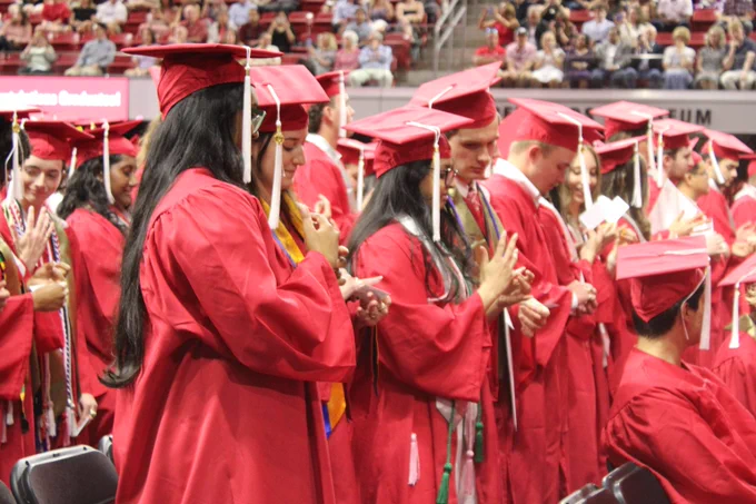 Graduates in red caps and gowns stand in rows during a commencement ceremony inside a large venue filled with spectators.