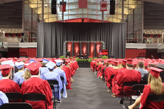 Graduates in red and light blue caps and gowns seated facing a stage with a gray curtain backdrop in a large indoor venue.