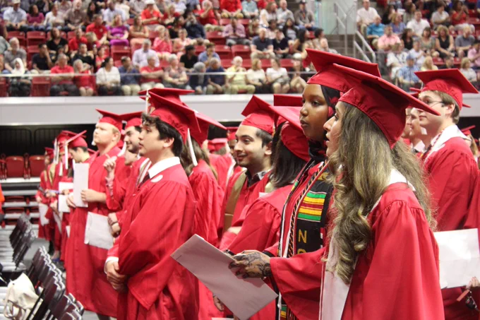 Graduates in red caps and gowns stand in rows during a ceremony with a large audience in the background.
