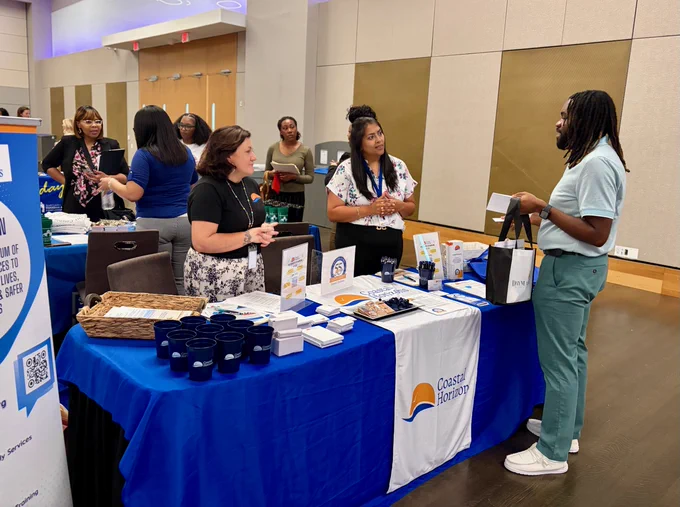 A group of people are interacting at an indoor event with various tables showcasing information and merchandise. A table prominently displays a banner with the name Coastal Horizons.