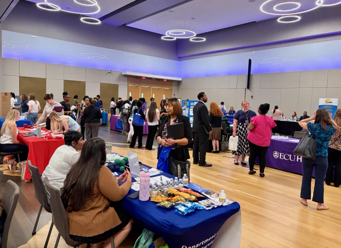 A large indoor career fair with multiple booths featuring organizations. People are engaged in conversations, and tables have informational materials and snacks.