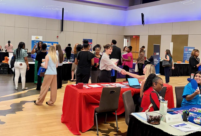 A career fair taking place in a spacious hall with several booths. Attendees interact with representatives at tables.  Tables are covered with promotional materials, and some people are seated while others stand and engage in discussions.