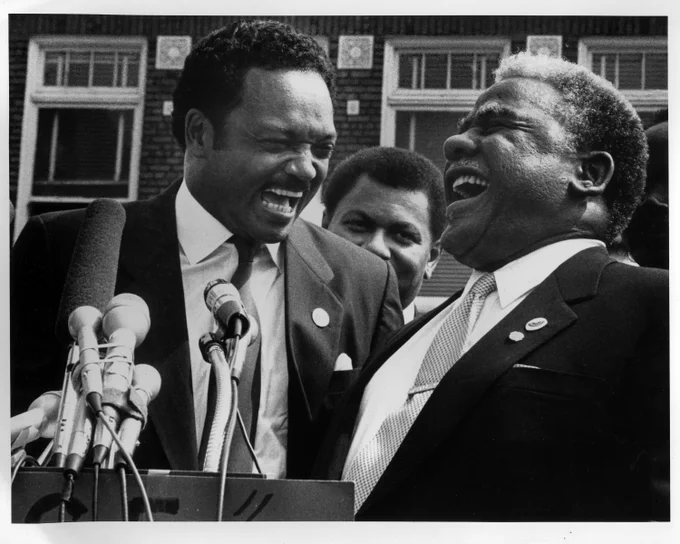 Black and white Photo of Mayor Harold Washington and Reverend Jesse Jackson, Sr. standing at a news podium and laughing together.