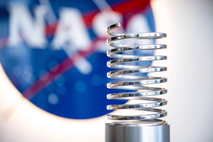 A Webby trophy, which is a silver spring spiraling up from a solid silver base. It sits in front of an out of focus NASA meatball logo, a blue circle with a red V and white loop, with NASA in white.