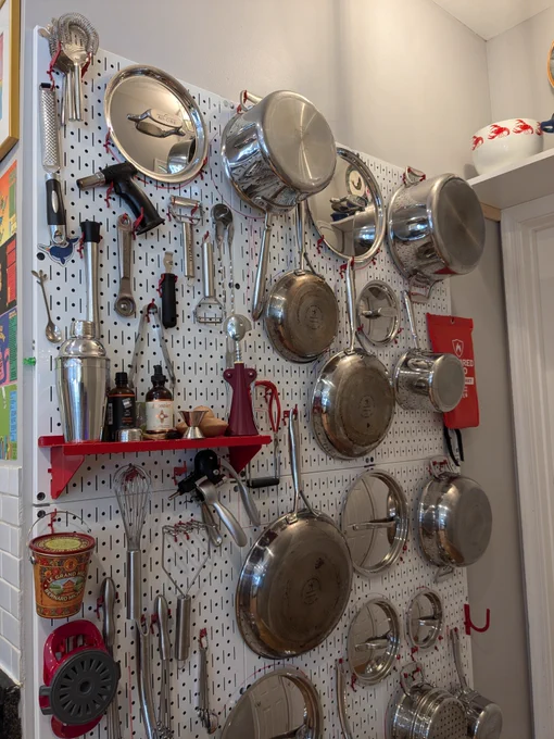 Alt text: A wall-mounted white metal pegboard system displays an organized arrangement of stainless steel pots, pans, lids, and kitchen tools. Items are neatly hung using hooks and accessories, with a small red shelf holding bottles and bar tools. The setup maximizes vertical space while creating a clean, functional, and visually appealing kitchen storage solution.
